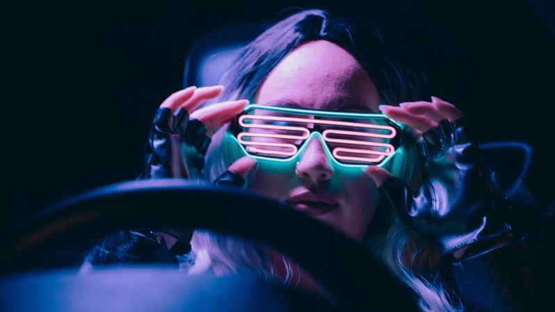 Dziewczyna siedzi za kierownicą, rękami sięga do neonowych okularów, które ma na nosie