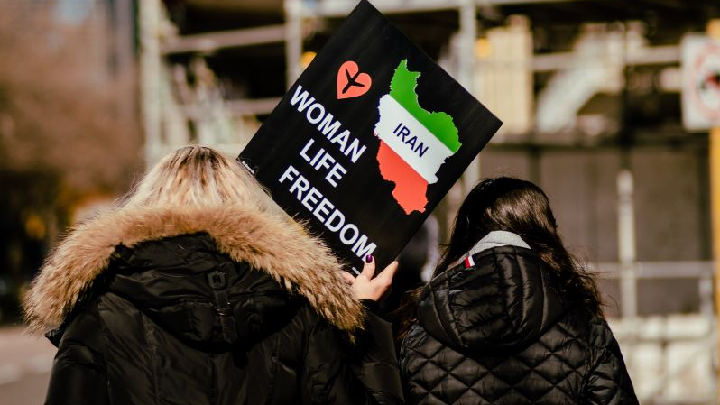 Dwie kobiety widziane z tyłu, trzymające transparent z hasłem dot. walki o prawa kobiet w Iranie